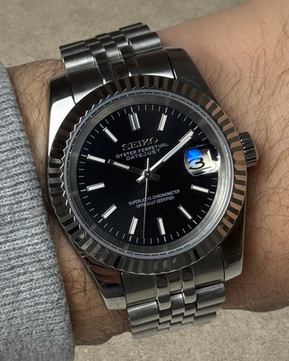 PB TimePiece Classic Date watch with a black dial, fluted bezel, and stainless steel jubilee bracelet on a wrist.