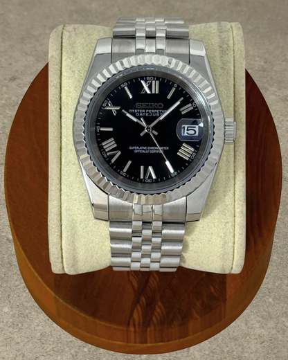 PB TimePiece Classic Date watch with a black dial, Roman numerals, fluted bezel, and stainless steel Jubilee bracelet.