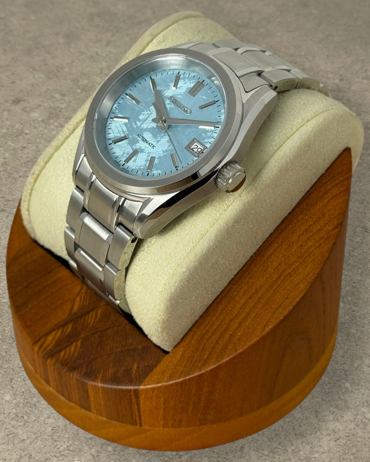 PB TimePiece Heritage watch with an ice blue textured dial, stainless steel case, and bracelet on a wooden stand.