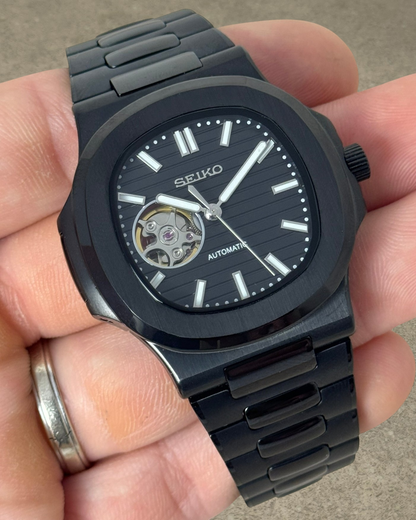 PB TimePiece Novara automatic watch with black stainless steel case, bracelet, and open heart dial detail.