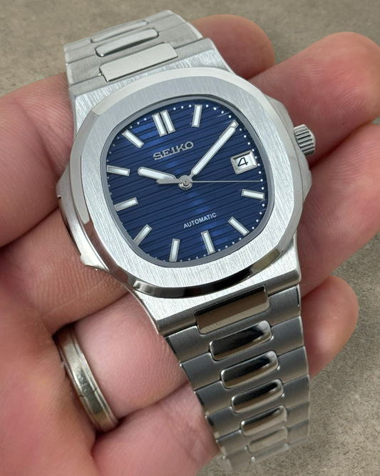PB TimePiece Novara automatic watch with a textured blue dial, stainless steel case, and brushed bracelet.