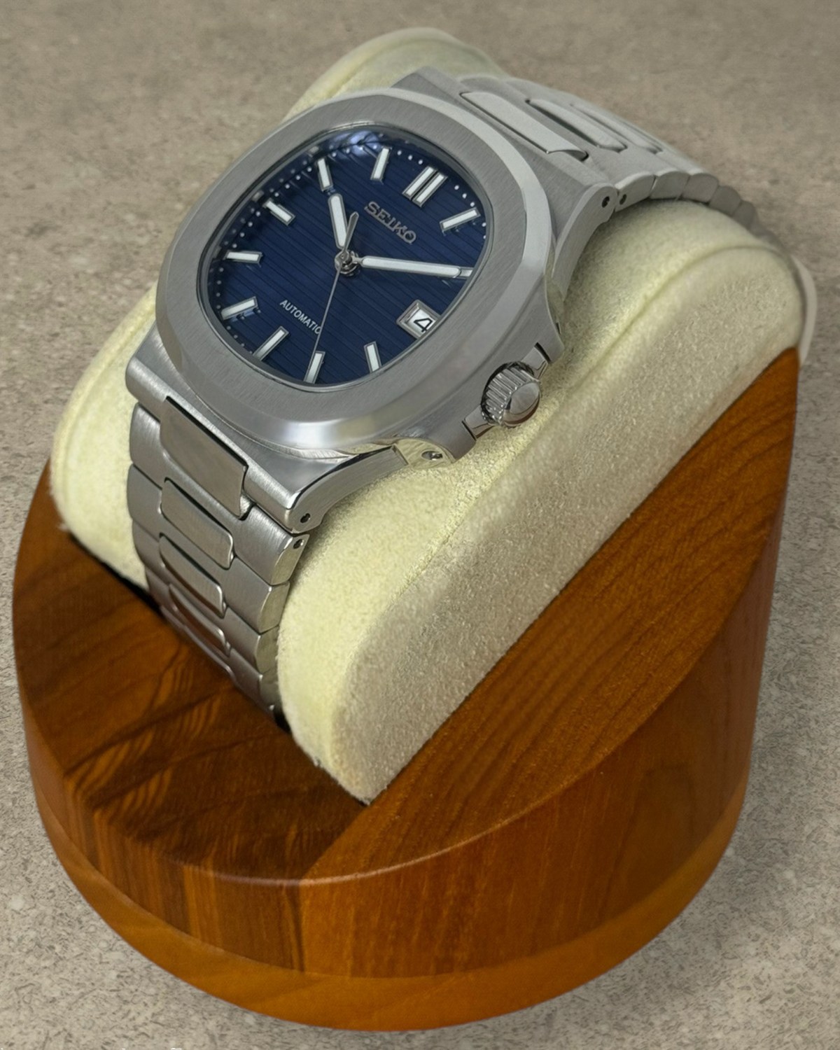PB TimePiece Novara watch with a textured blue dial, stainless steel case, and bracelet on a wooden display stand.