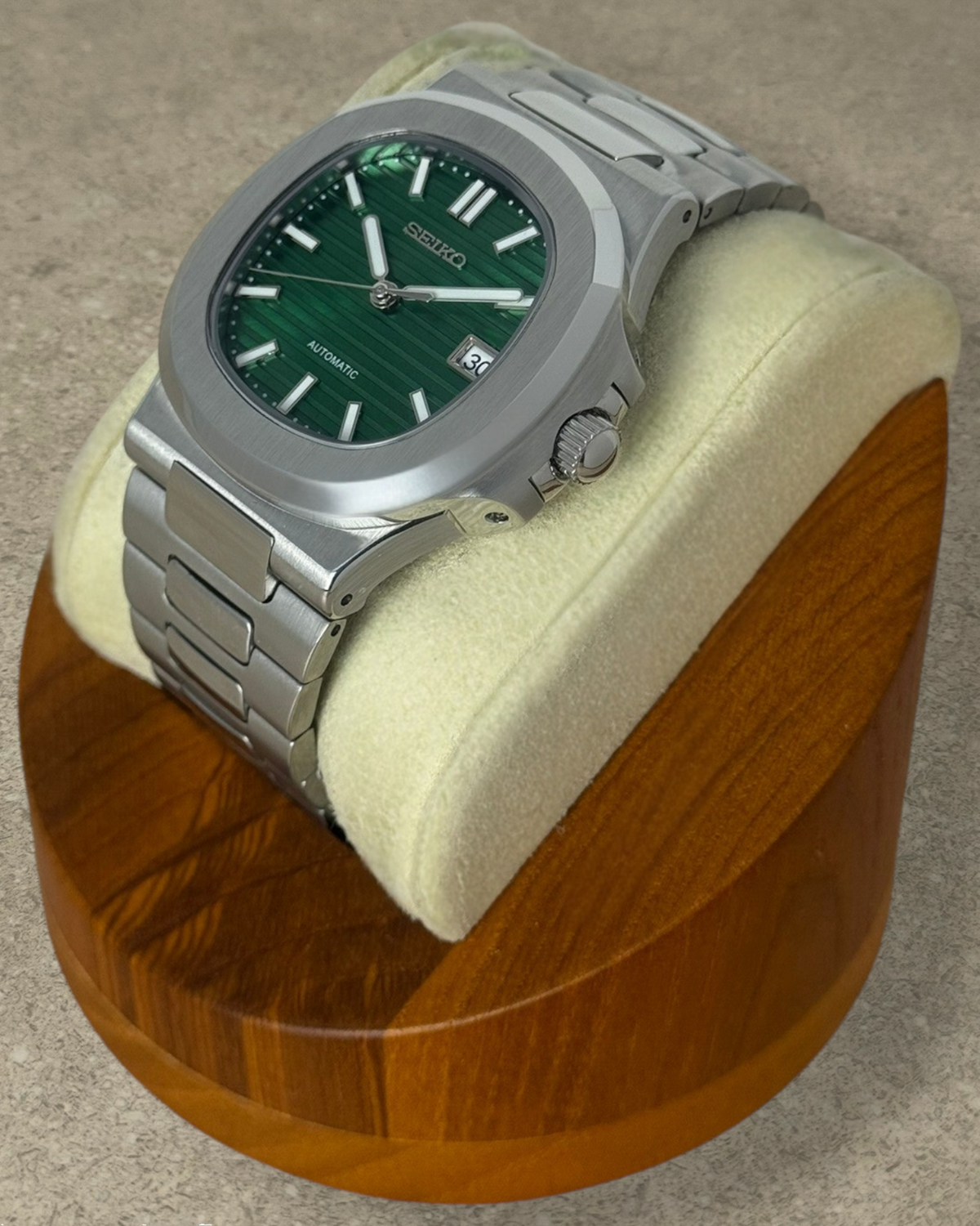 PB TimePiece Novara watch with a textured emerald green dial, stainless steel case, and bracelet on a wooden stand.