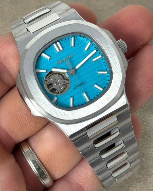 PB TimePiece Novara automatic watch with a teal blue open heart dial, stainless steel case, and brushed bracelet.