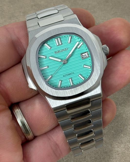 PB TimePiece Novara watch with a textured teal dial, stainless steel case, and bracelet held in a hand.