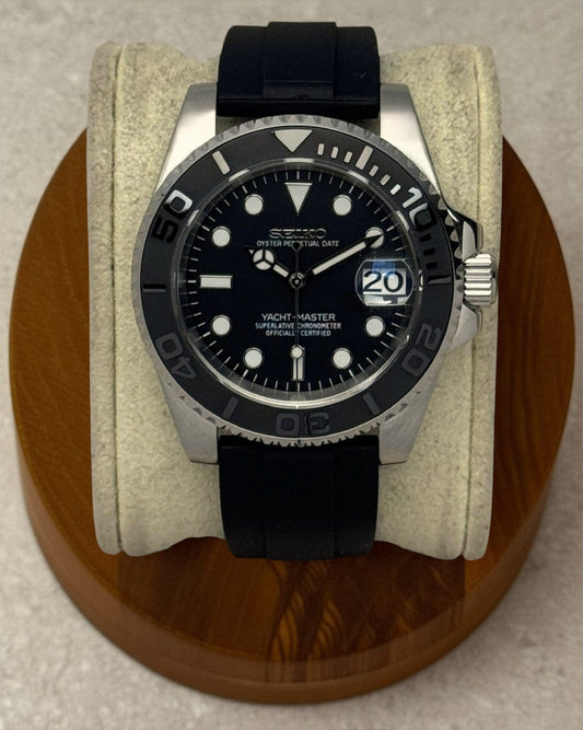 PB TimePiece Yachtmaster watch with a matte black dial, rotating bezel, and black rubber strap on a wooden stand.